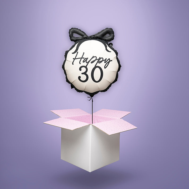Foliowy balon z helem w kształcie okrągłym, beżowy z czarnym napisem ‘Happy 30’ i ozdobną czarną kokardą, unoszący się nad otwartym pudełkiem z różową bibułką. Balonowa niespodzianka wysyłka cała Polska.