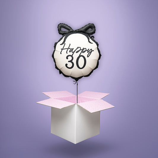 Foliowy balon z helem w kształcie okrągłym, beżowy z czarnym napisem ‘Happy 30’ i ozdobną czarną kokardą, unoszący się nad otwartym pudełkiem z różową bibułką. Balonowa niespodzianka wysyłka cała Polska.