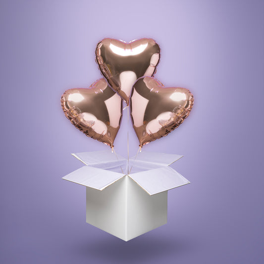 Trzy balony foliowe w kształcie serc w odcieniu rose gold, wypełnione helem i wyskakujące z białego kartonu – elegancka niespodzianka na Walentynki, rocznicę lub ślub.