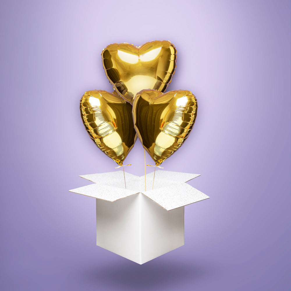 Trzy złote balony foliowe w kształcie serc, wypełnione helem i wyskakujące z białego kartonu – elegancka niespodzianka na Walentynki, rocznicę lub ślub.